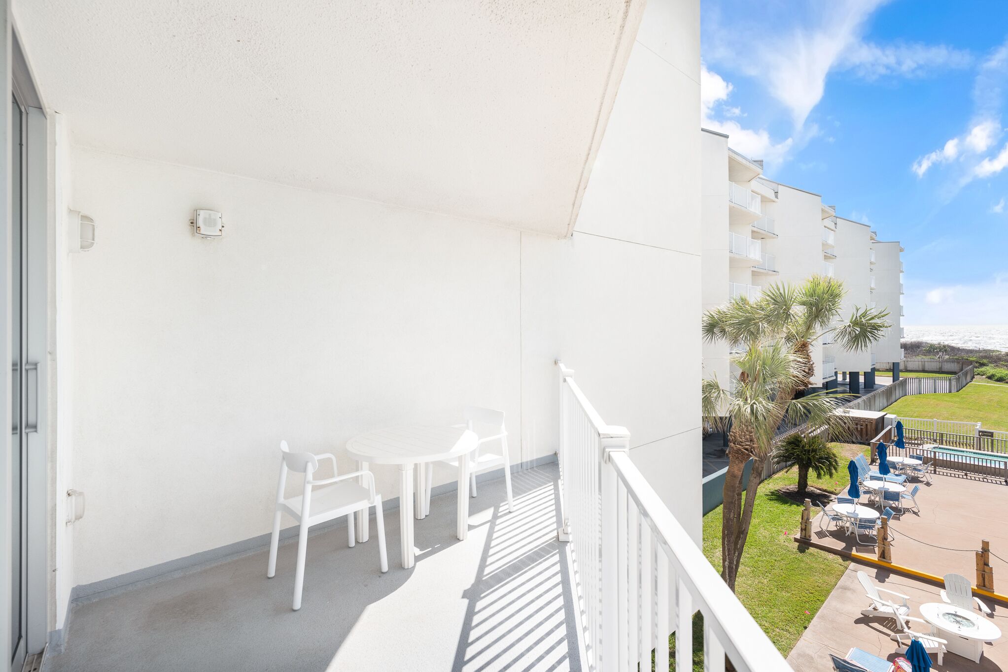 A balcony with white chairs and a table, overlooking a pool area with lounge chairs and palm trees.