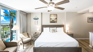A bedroom with a large bed, a ceiling fan, a nightstand with a lamp, a chair, and a view of the ocean through a balcony. - Sandcastle Condominiums Unit 217 (Port Aransas)