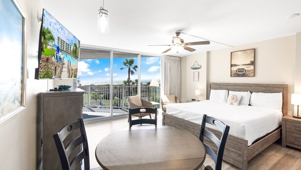 A hotel room with a bed, a dining table, chairs, a ceiling fan, a large window with a view of palm trees, and a painting on the wall. - Sandcastle Condominiums Unit 217 (Port Aransas)