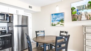 A modern kitchen with stainless steel appliances, a dining table with four chairs, and a beach-themed painting on the wall. - Sandcastle Condominiums Unit 217 (Port Aransas)