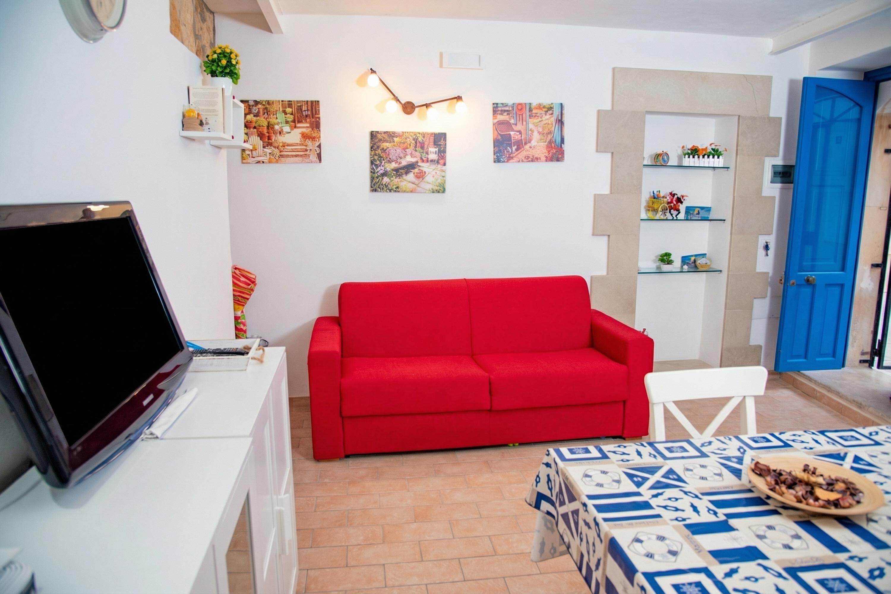 Ein Wohnzimmer mit einem roten Sofa, einem Fernseher auf einem weißen Schrank, einem gemusterten Tisch und einer blauen Tür.