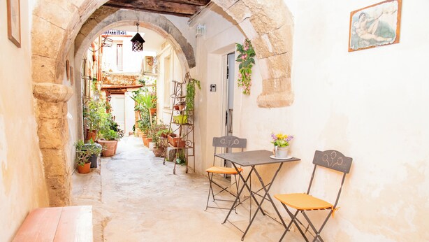 Un estrecho callejón con una mesa, sillas, plantas en macetas y un banco.