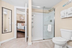 A modern bathroom with a glass-enclosed shower, a white toilet, and a visible bedroom with a bed and a mirror.