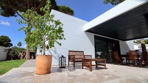 A modern outdoor seating area with a large potted tree, wooden chairs, and a glass door entrance. - Villa del Fresno with pool and parking (Chiclana)