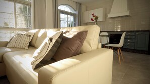 A modern living room with a beige sofa, a dining area with a white chair, and a kitchen with blue cabinets. - Hibiscus Apartment with Shared Pool (Chiclana de la Frontera)