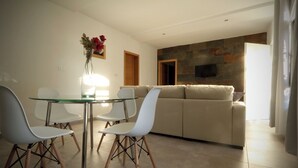 A modern living room with a glass dining table, white chairs, a beige sofa, and a stone accent wall. - Hibiscus Apartment with Shared Pool (Chiclana de la Frontera)