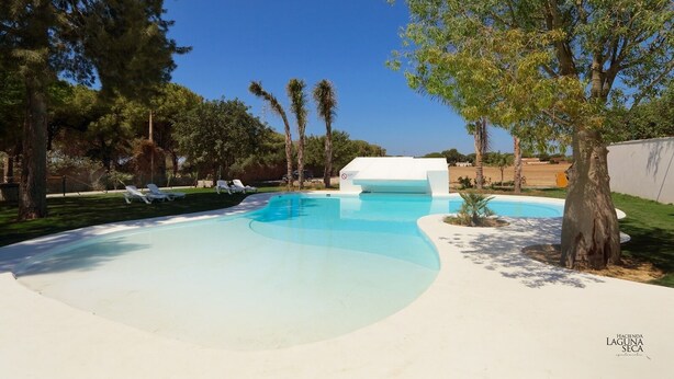 A swimming pool with lounge chairs, surrounded by trees and a white building. - Buganvilla Apartment with Shared Pool (Chiclana de la Frontera)
