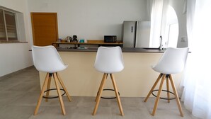 A kitchen with three white bar stools, a wooden door, a refrigerator, and a window with curtains. - Buganvilla Apartment with Shared Pool (Chiclana de la Frontera)
