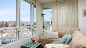 Uma sala de estar com sofá, uma mesa pequena com um vaso de flores e vista para prédios através de grandes janelas.