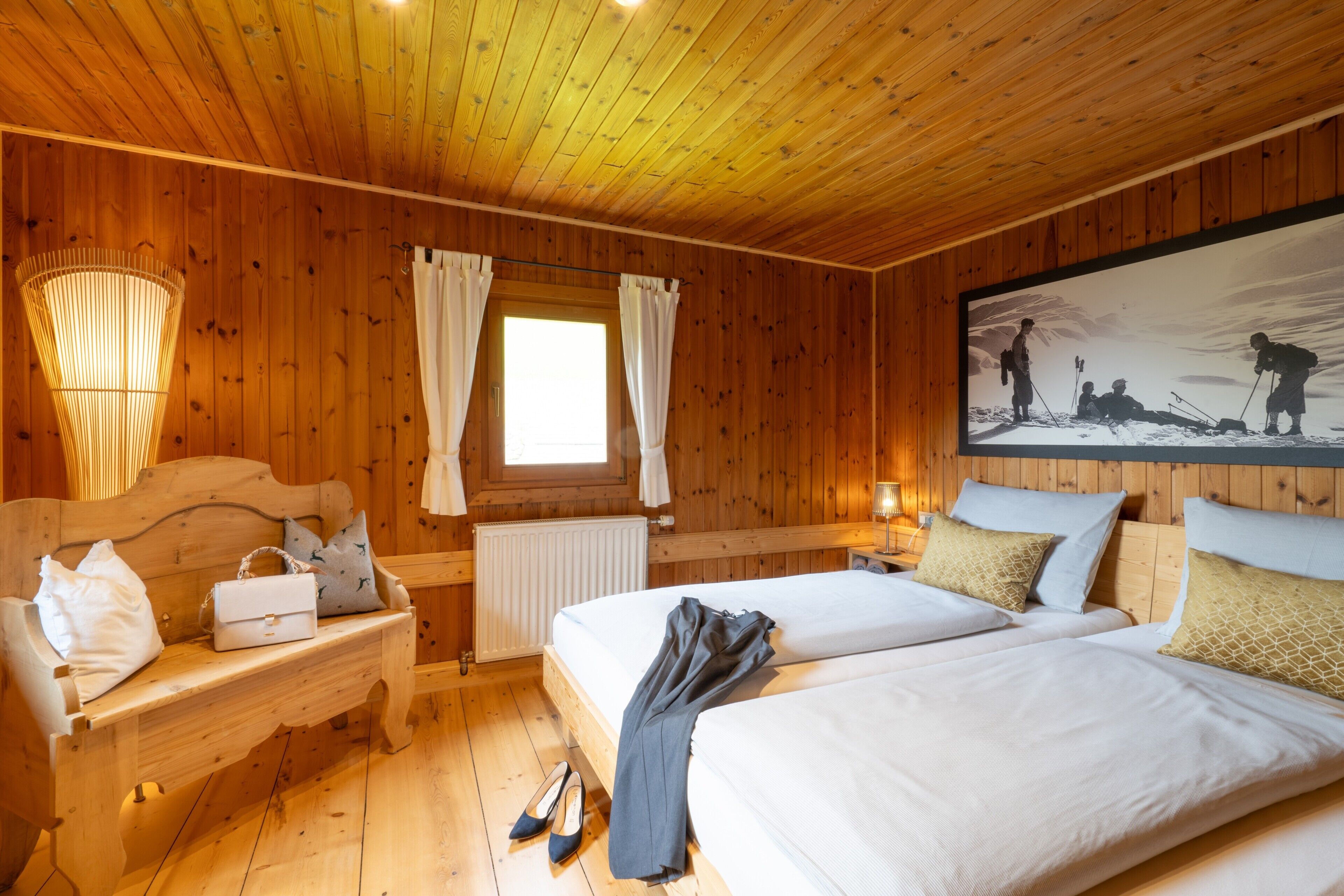 Una habitación con paneles de madera, dos camas, un banco con bolsos, un radiador y un cuadro en la pared.