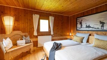 Una habitación con paneles de madera, dos camas, un banco con bolsos, un radiador y un cuadro en la pared.