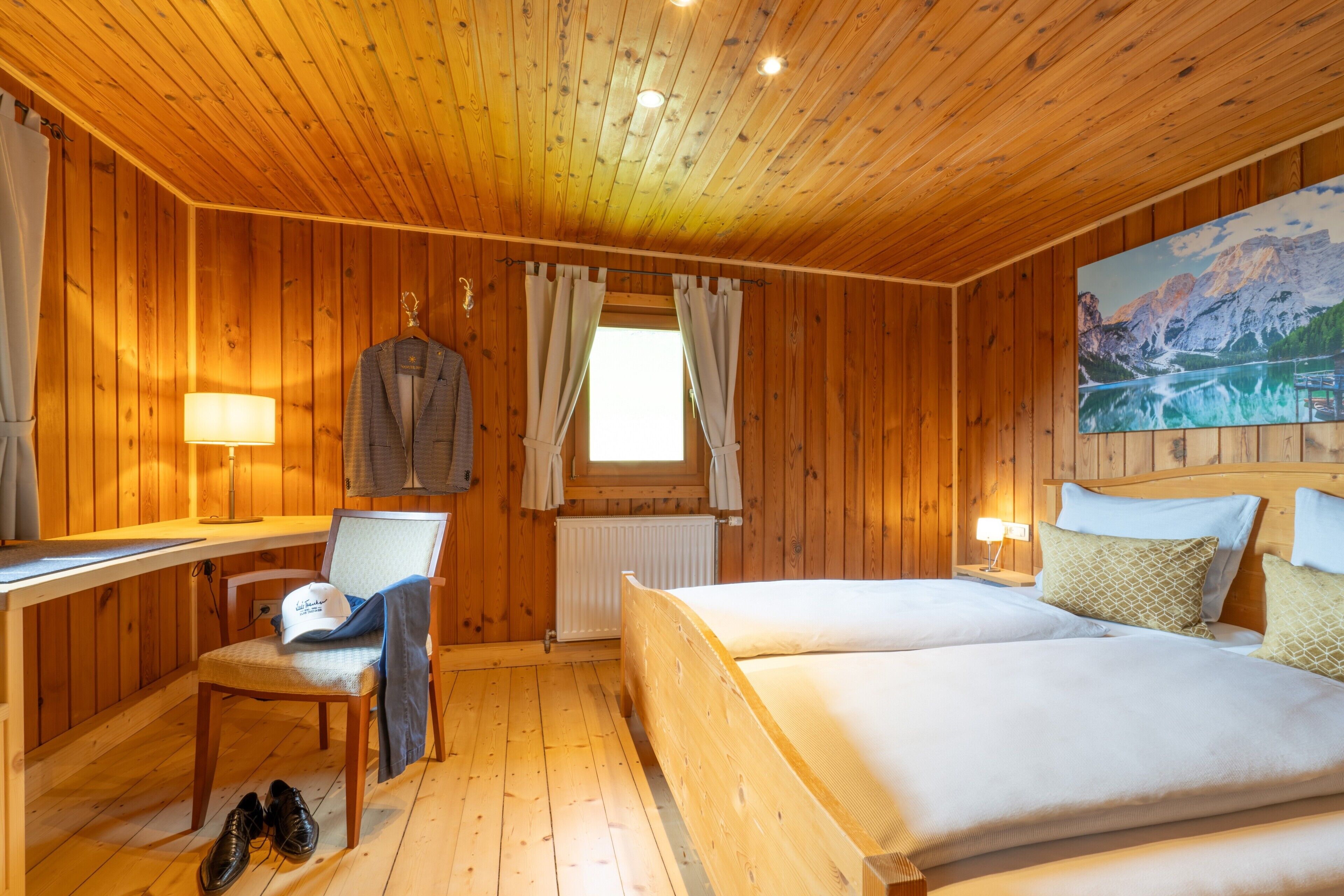 A wooden cabin room with a bed, a desk, a chair, and a window with curtains.
