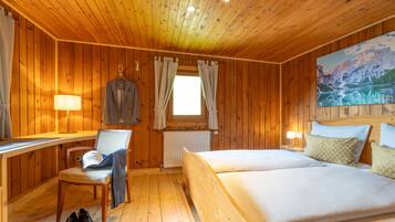 A wooden cabin room with a bed, a desk, a chair, and a window with curtains.