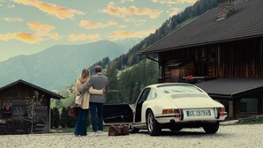 A couple standing by a white car with a license plate GE 287BA in a rural setting with wooden buildings and mountains in the background.