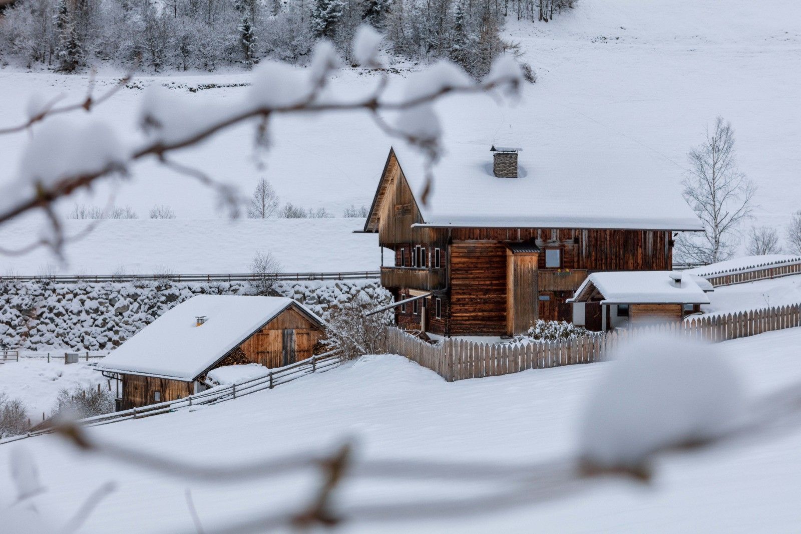 Una cabaña de madera con techo pronunciado y una estructura pequeña adosada, rodeada por un paisaje nevado.