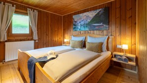 A wooden room with a bed, a window with curtains, a painting, and a nightstand with a lamp.