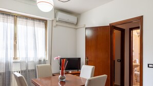 Un comedor con una mesa de madera, sillas blancas, un jarrón con flores, un televisor y un equipo de aire acondicionado.