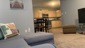 A living room with a gray sofa, a brown ottoman, and a TV stand. A kitchen is visible in the background with wooden cabinets and a microwave. - Cozy Basement Suite close to Downtown (Edmonton)