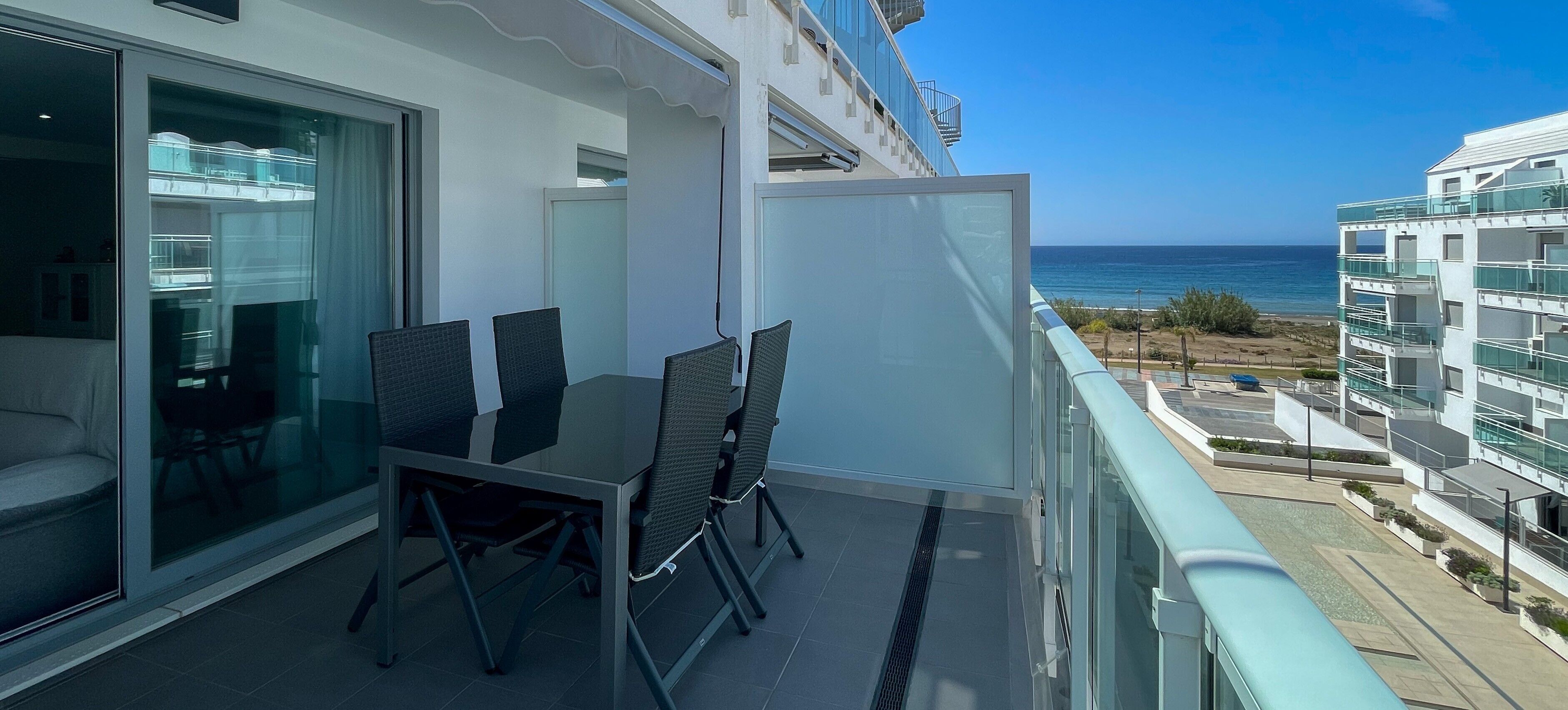 A balcony with a table and chairs, a glass partition, and a view of the ocean and buildings.