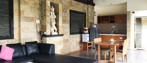Una sala de estar con un sofá negro, una mesa de comedor con sillas, una zona de cocina con armarios de madera y una pared de piedra con una estatua.