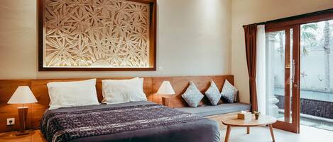 Quarto de hotel com uma cama grande, cabeceira de madeira, painel decorativo na parede, mesa de cabeceira com abajur e vista para o exterior através de uma porta de vidro.