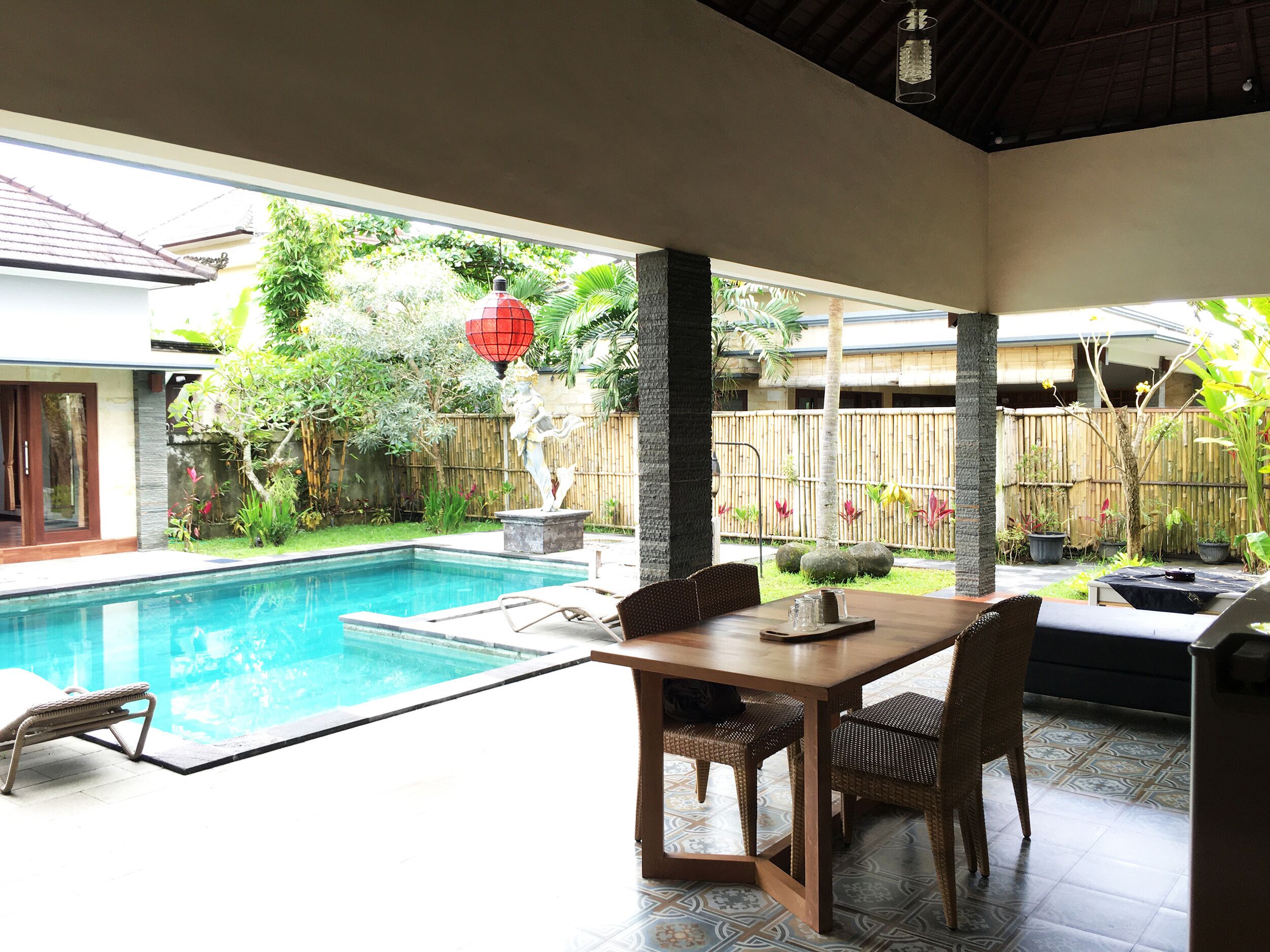 A poolside area with a dining table and chairs, a statue, and a view of a bamboo fence and greenery.