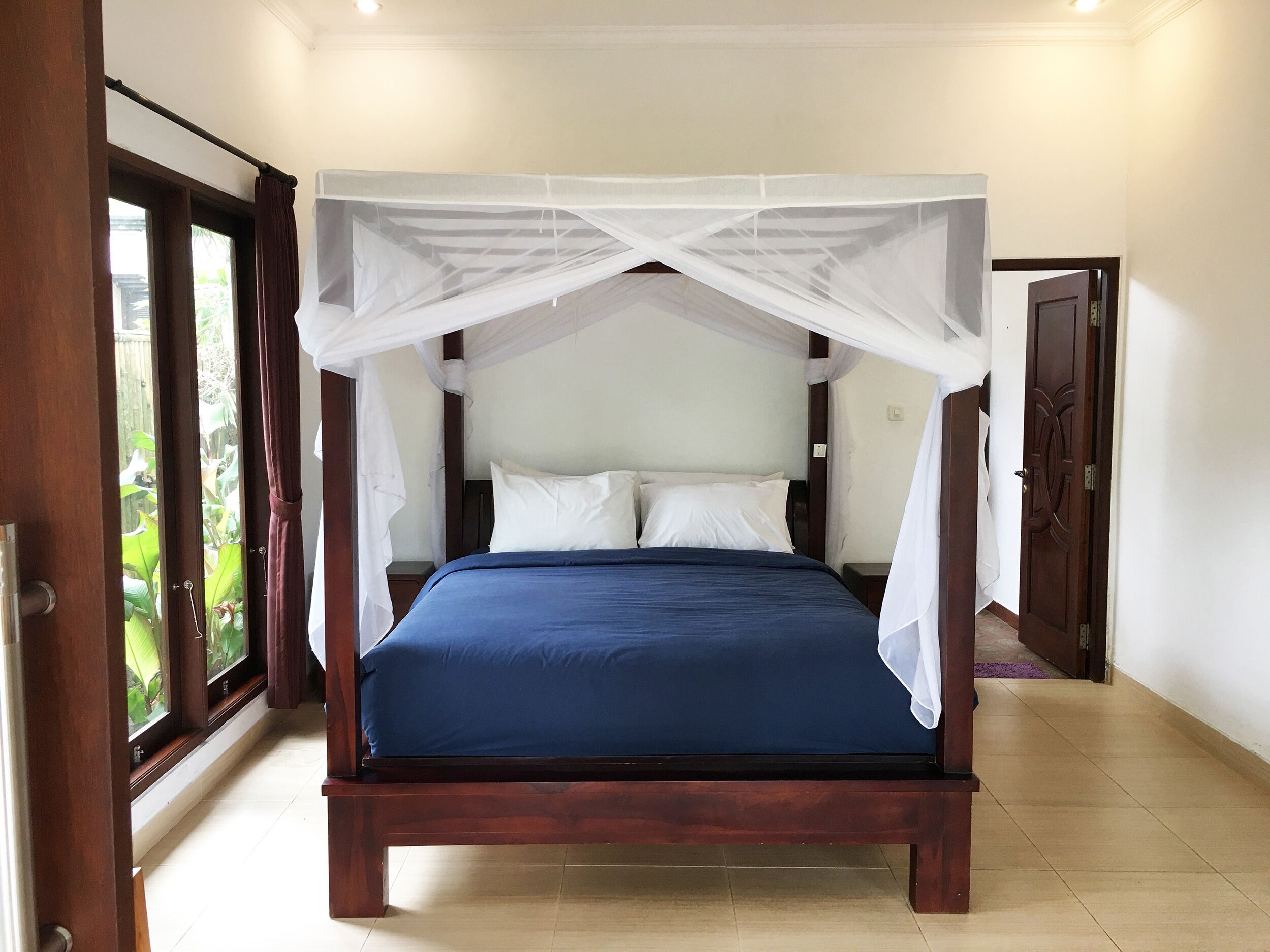 A bedroom with a four-poster canopy bed, a wooden headboard, and a view of greenery through the window.