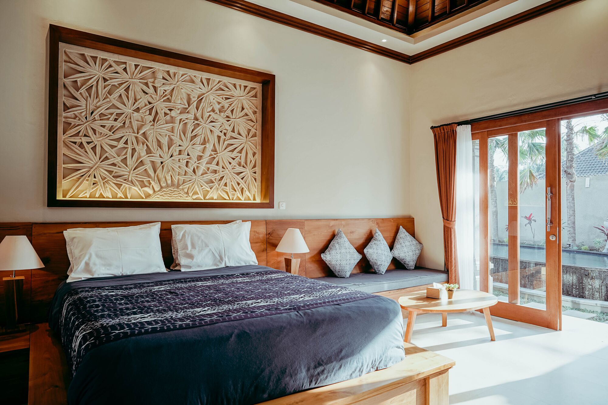 Un dormitorio con una cama grande, cabecera de madera, una obra de arte en la pared, una mesita de noche con lámpara y vistas al exterior a través de una puerta corrediza.