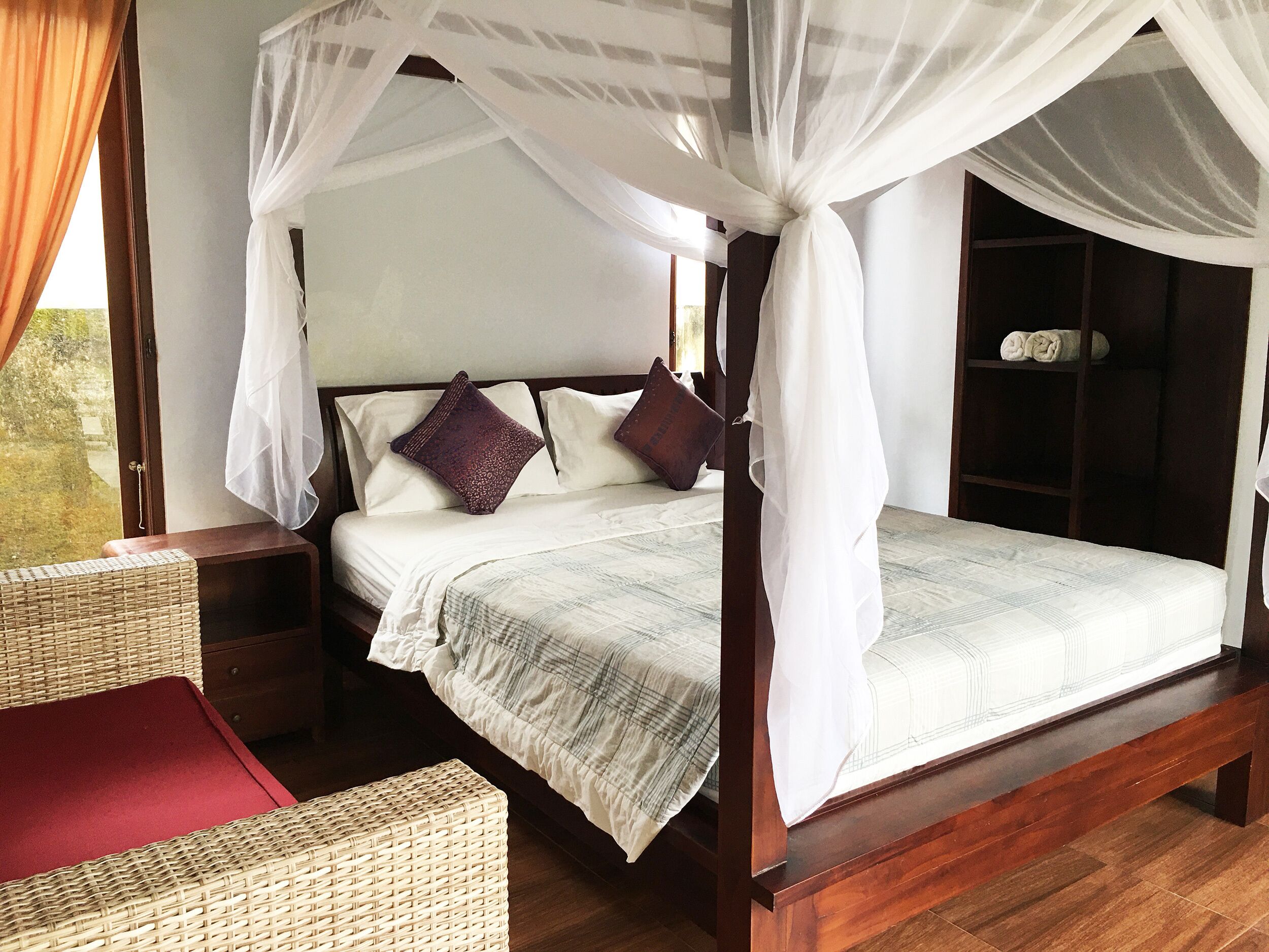 A four-poster canopy bed with a wooden frame, white bedding, and a view of the outdoors through a window.