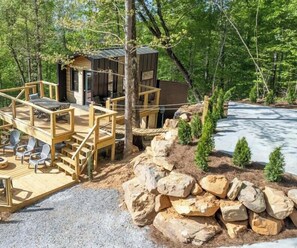 A cabin with a wooden deck and outdoor seating, surrounded by trees and a rocky landscape. - The Lumber Jack Pine Treehouse at Treetop Escapes! (Wildwood)
