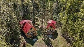 Deux cabanes en bois aux toits rouges, entourées d’une forêt dense, chacune dotée d’un petit porche et d’un escalier apparent menant à l’entrée.
