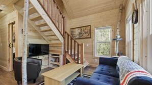 L’intérieur d’un chalet en bois, avec un escalier, un canapé, un téléviseur et une fenêtre donnant sur les arbres.