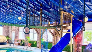 Zona de piscina interior con un tobogán azul, red de cuerdas y una cubierta de madera.