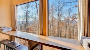 Una habitación con un amplio ventanal con vistas a un bosque, un banco de madera y una pequeña mesa con toallas.