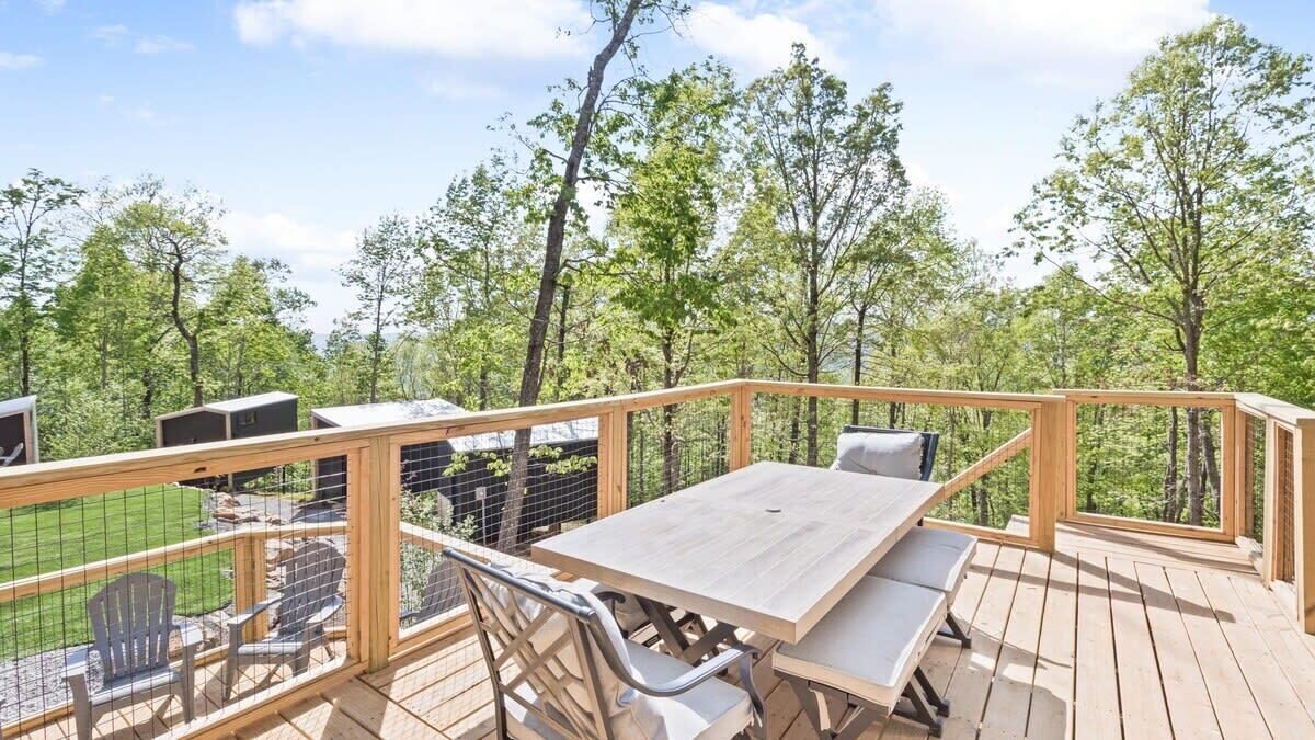 A wooden deck with a table and chairs, surrounded by a wooden railing, overlooking a green landscape with trees.