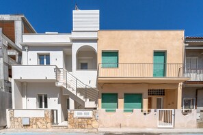 Un edificio de varios pisos con fachada de piedra, un balcón con contraventanas verdes y una escalera que conduce a un nivel superior.