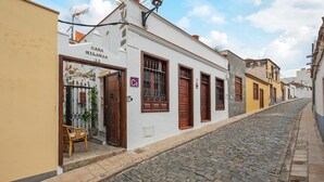 Une ruelle étroite pavée, bordée d’un immeuble blanc portant l’inscription « CASA HILARIA 13 », avec une devanture de magasin dotée d’une porte en bois et de volets aux fenêtres.