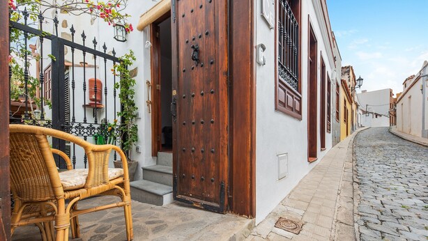 Une rue pavée, une porte en bois, une chaise en osier et un bâtiment blanc avec une clôture en fer noir.