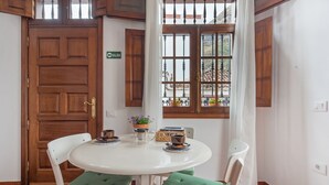 Un coin repas avec une table ronde, deux chaises dotées de coussins verts, une cafetière, une petite plante et une fenêtre donnant sur un immeuble.
