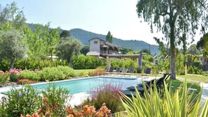 Zona de piscina con sillones de descanso, rodeada de exuberante vegetación y un edificio al fondo.