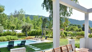 Zona de piscina con sillones de descanso, una mesa de comedor con sillas y vistas a un jardín exuberante y colinas al fondo.