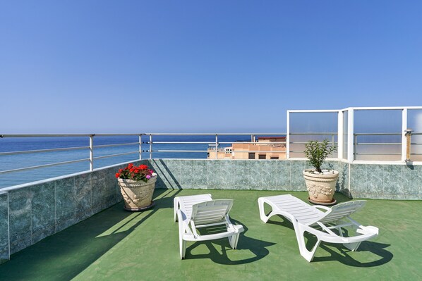 A rooftop terrace with two white lounge chairs, a potted plant with red flowers, and a view of the sea.