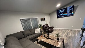 A modern living room with a sectional sofa, a coffee table with decorative items, a leather chair, and a wall-mounted TV displaying a Pink Floyd playlist.