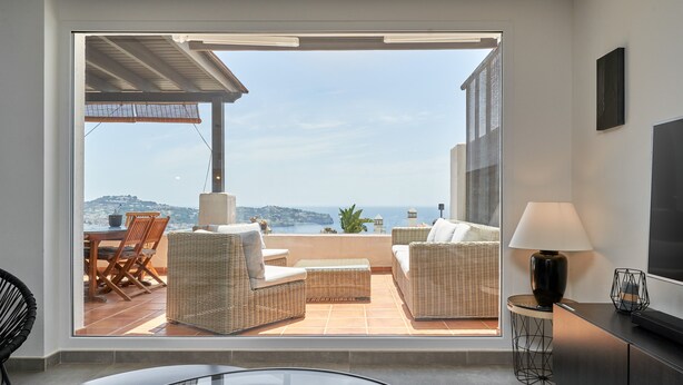 Un salon moderne avec une porte vitrée donnant sur un balcon aménagé avec des meubles en osier et offrant une vue sur la mer.