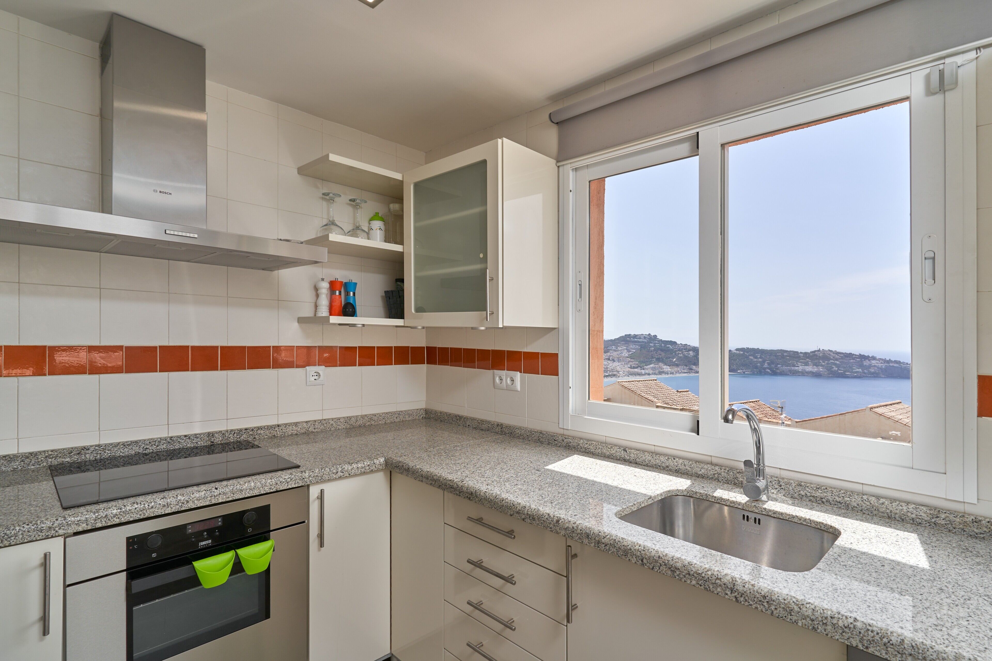 A kitchen with a built-in oven, a stainless steel range hood, a window with a view of the sea, and a granite countertop.
