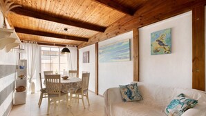 Een kamer met een houten plafond, een eethoek met een tafel en stoelen, een witte bank met decoratieve kussens en kunstwerken aan de muren.