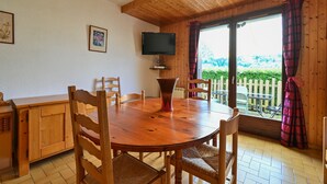 Una mesa de madera con sillas, un televisor montado en la pared y vistas a la vegetación exterior.