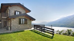 Una casa de piedra con balcón, cerca de madera y vista a un lago y montañas.