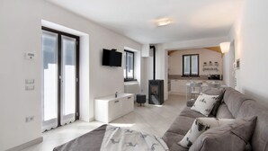 Una sala de estar moderna con un sofá gris, un mueble blanco, un televisor en la pared y una cocina al fondo.