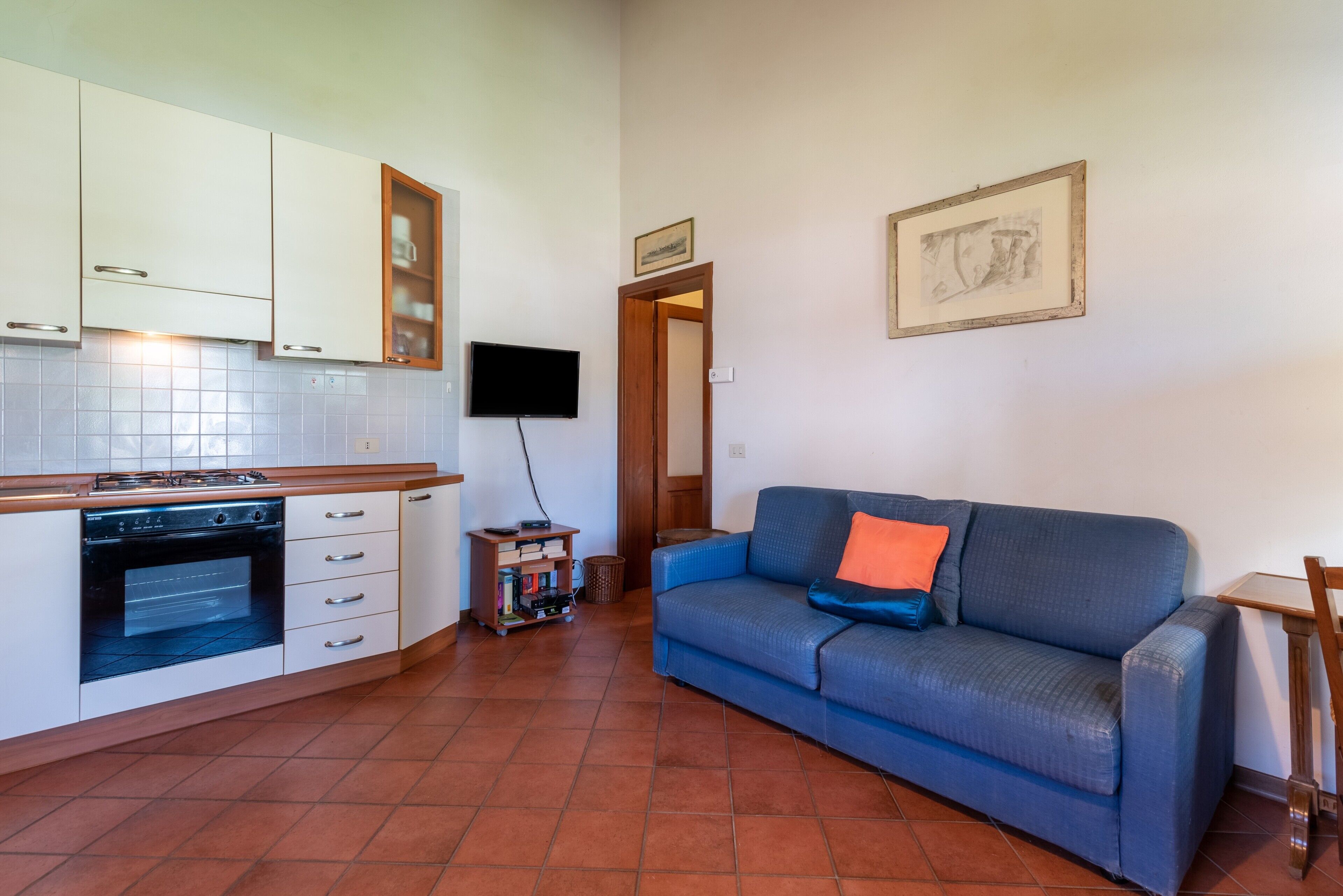 A living room with a blue sofa, a television, and a kitchen area with white cabinets and a tiled backsplash.